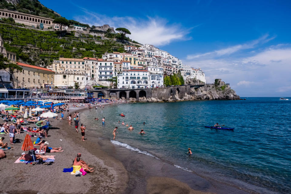 A beach with people sunbathing and swimming, set against a hillside town with terraced buildings and a clear blue sky