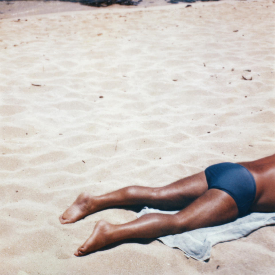 Person sunbathing on a sandy beach, lying on a towel, wearing swim trunks. No famous individuals identifiable