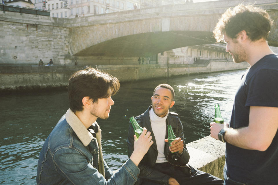 Three friends sit by a river, holding bottles, and chatting under a bridge on a sunny day