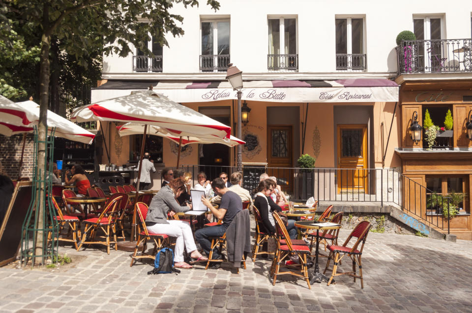 People dining outdoors at a Parisian café terrace with red chairs, under umbrellas, set against a classic café façade