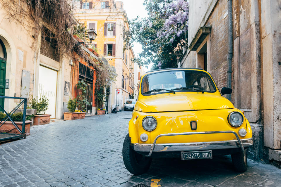 A yellow vintage car parked on a cobblestone street in a European city with rustic buildings and greenery