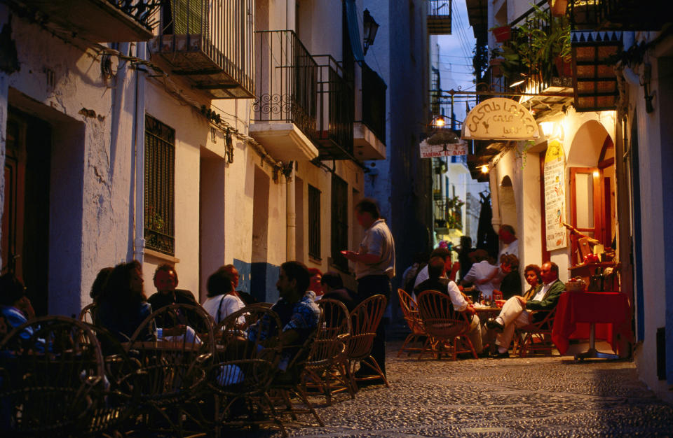 A bustling outdoor café scene in a narrow, cobblestone street, with people dining and socializing in the evening