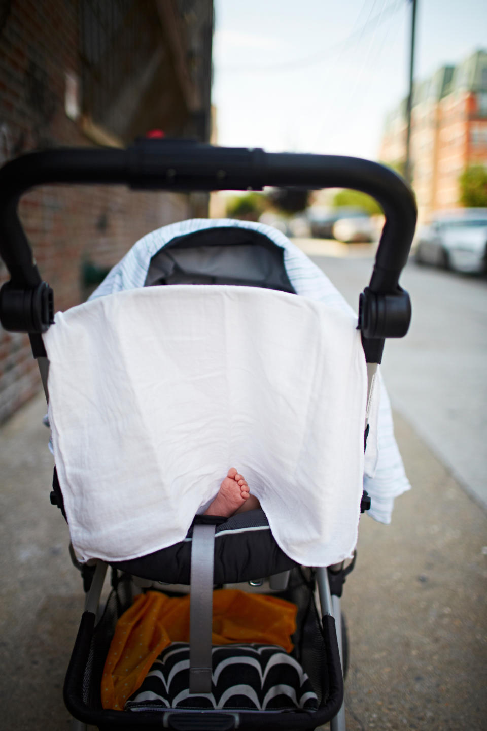 Baby's tiny foot peeks out from under a white cloth draped over a stroller, on an urban sidewalk