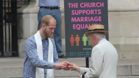 Pastor Steve Ames Pastor Steve Ames, who is wearing a blue and white paisley print button shirt with a white stole over his neck, hands an older man wearing a flat top hat and light coloured suit jacket a small red cup. They are stood outside a white stone building beside a sign that says 'The church supports marriage equality'.