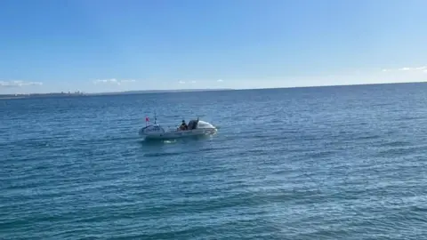 Sway PR Zara Lachlan in a white wide-beamed 24ft (7m)-long boat. She is rowing on blue ocean water, which is calm, with paler blue sky above