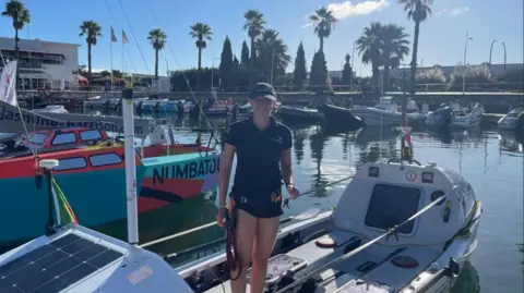 Sway PR Zara Lachlan wearing her hair tucked into a dark blue baseball cap and a dark blue polo shirt, both with Team Forces and the Union flag logos. She is standing in a white boat just before she set off on the challenge. It is in a marina with other boats around it.