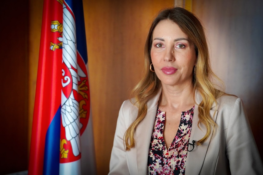 A woman looks at the camera with a neutral expression. In the background, a Serbian flag is visible.