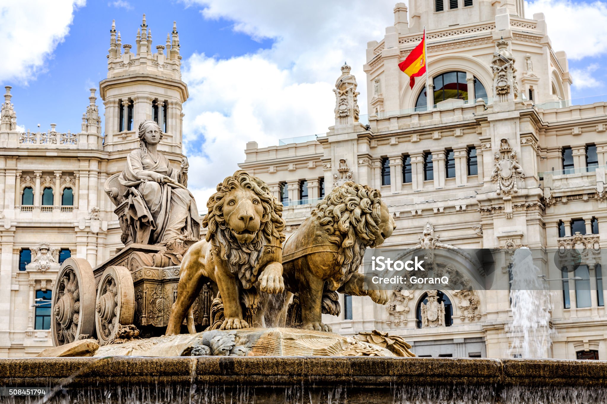 The Cibeles Fountain in Madrid features a statue of the goddess Cybele on a chariot pulled by lions, with a historic building and Spanish flag in the background.