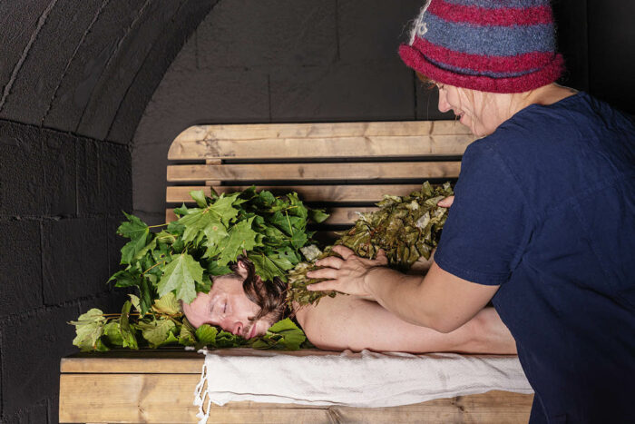 In a wooden room, a person uses a bunch of small tree branches to pat the back of a person lying on a bench.