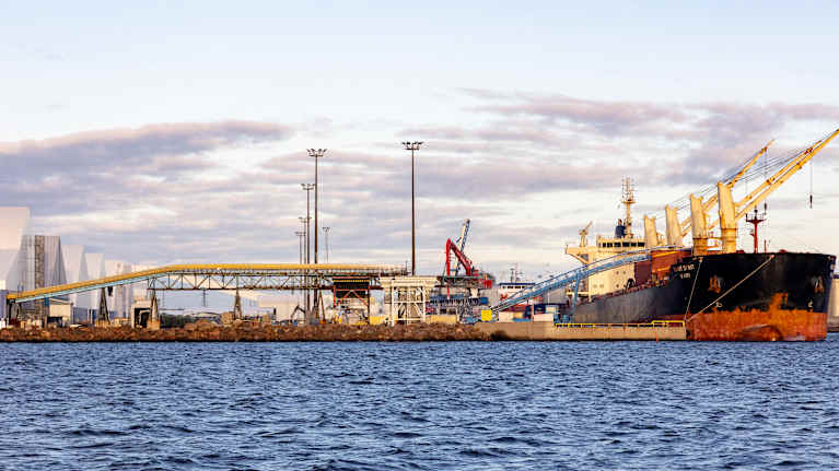 A large rusty black ship docked in a port with cranes and buildings. 