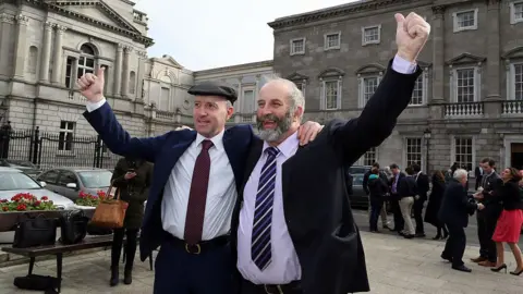 Getty Images Michael Healy Rae (left) with his brother Danny Healy-Rae (right). Both men are wearing suits and have an arm held in the air. Michael is wearing a hat. 