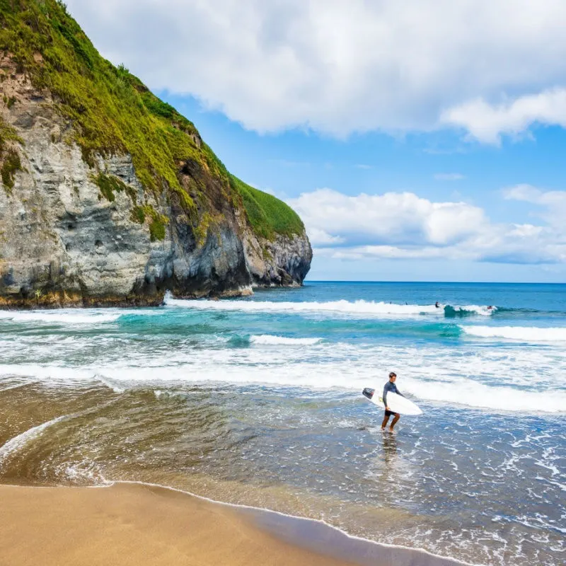 Man with surfing board walking in water across Santa Barbara beach, Sao Miguel island, Azores, Portugal.