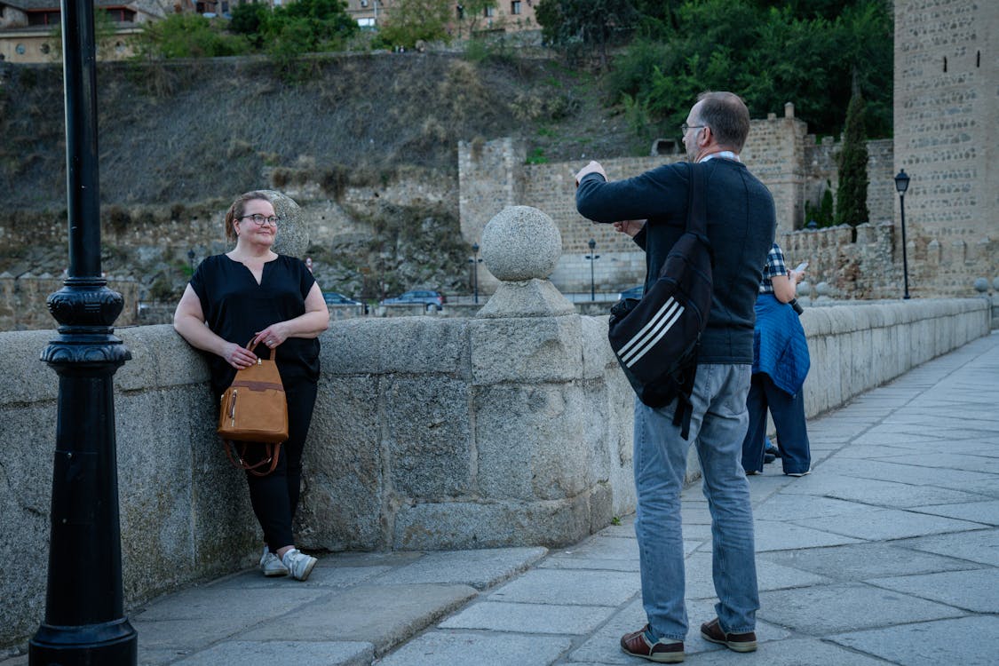 A person in black clothing poses against a stone wall while someone takes their photo on a historic street with ancient fortifications visible in background.