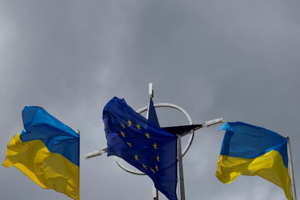 Two national flags of Ukraine flank the European Union flag in the foreground to the NATO emblem with storm clouds in the background in central Kyiv, Ukraine on July 11, 2023. REUTERS/Valentyn Ogirenko