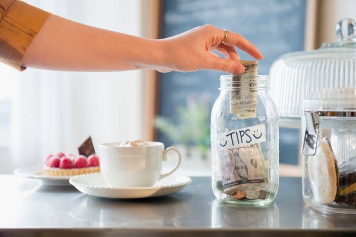 A hand placing money into a tip jar on a cafe counter with a coffee cup and pastry in the background