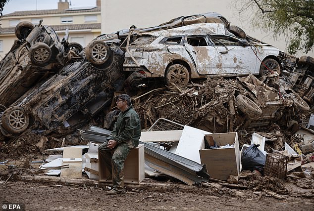 A volunteer rests next to a mountain of scrap metal and personal belongings piled up on a street in Paiporta