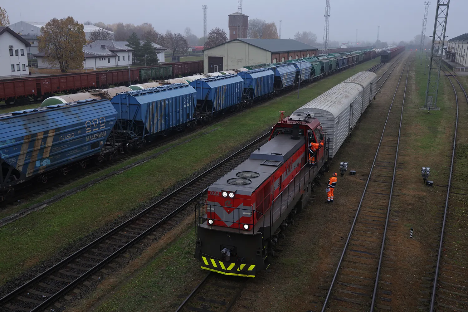 Two men in orange gear, one on a train, one on the ground, are at center on and next to a freight train. Another train stretches into the distance at left. Buildings are seen at left.