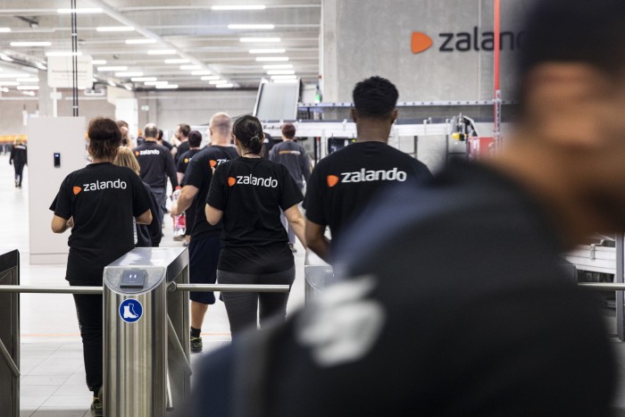 Workers dressed in matching Zalando-branded uniforms entering a large industrial workspace with visible equipment and organizational signage in the background