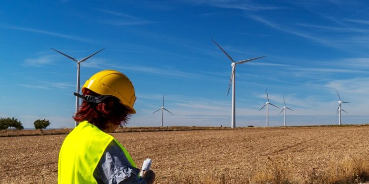 A woman dressed in a high-vis jacket and a hard hat stands in front of a field while turbines turn in the distance. It is a sunny day.