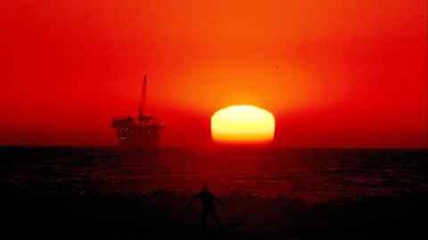 An oil rig out to sea seen from the shore, silhouetted against a red sky with the orb of a glowing sun dropping below the horizon