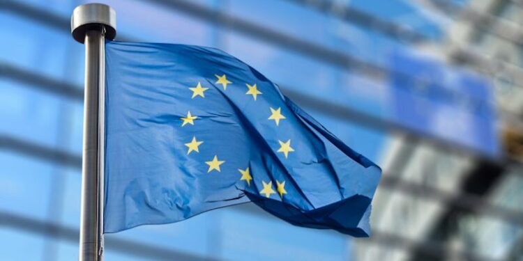 A waving blue flag adorned with a circle made up of stars flies outside of the official headquarters of the European Commission building.