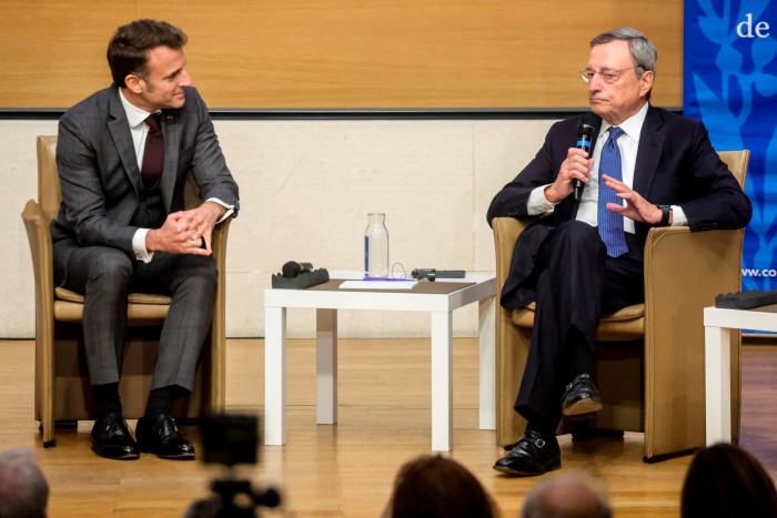 Two men in formal suits engaged in a discussion during a panel event. One is holding a microphone and speaking, while the other listens attentively