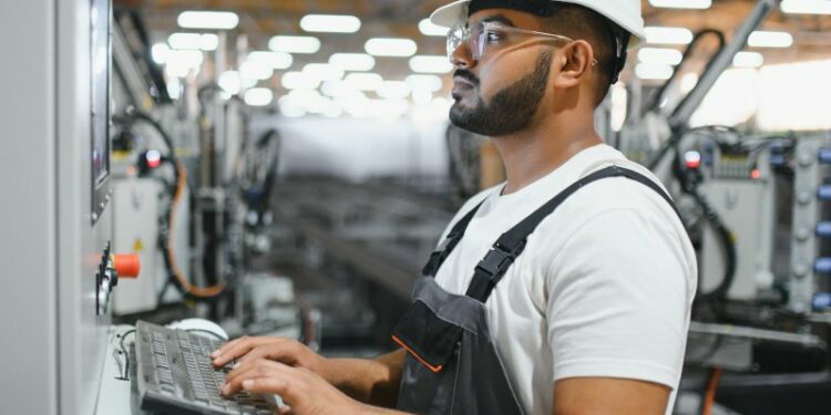 A young man wearing a hardhat and glasses operates a keyboard attached to some kind of technology console. He appears to be inside a factory building.