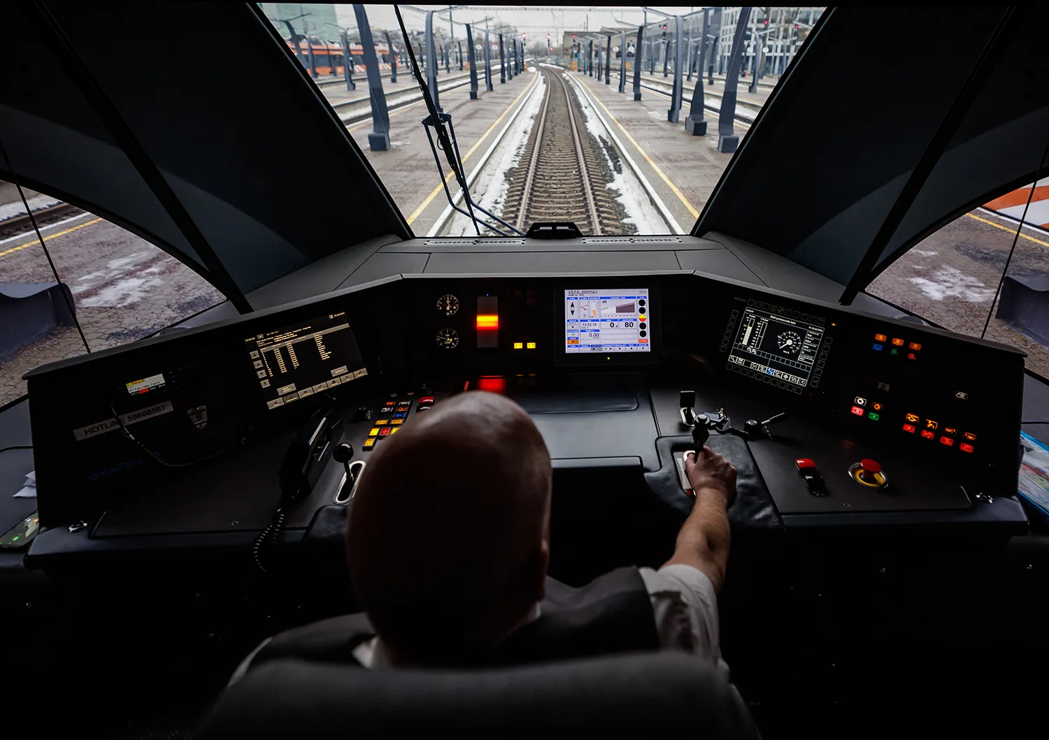 A conductor is seen from behind looking out down the track from the interior of a train at a train station