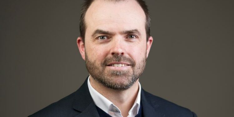 A man wearing a dark blazer over a jumper and white shirt smiles at the camera in front of a dark grey background.