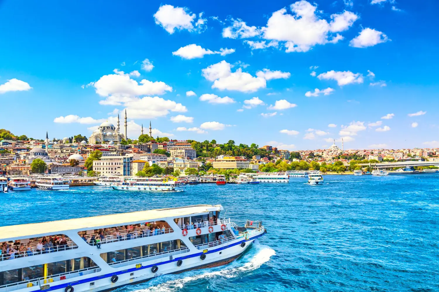 A scenic view of the Bosphorus in Istanbul, featuring a large passenger boat in the foreground and the historic skyline with mosques and colorful buildings in the background under a bright blue sky.