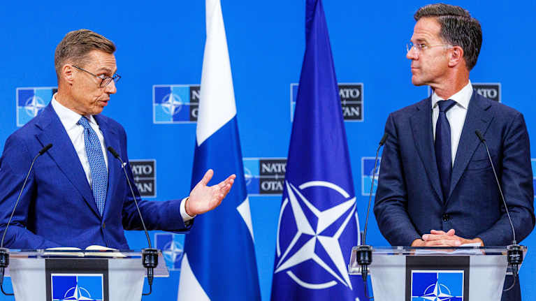 Two men in dark blue suits standing at podiums with the Nato logo and flag, as well as Finnish flag, in the background.