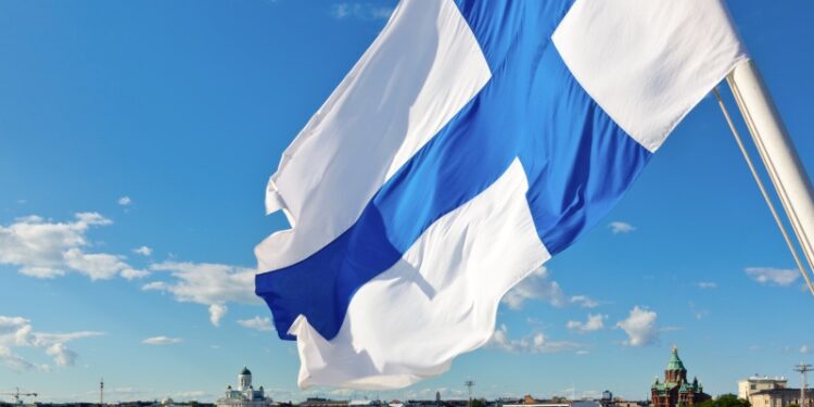 A Finnish flag blows in the wind. A city can be seen in the background.