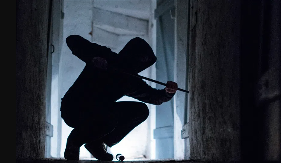 a hooded figure wearing black clothing using a crowbar to break into a door.