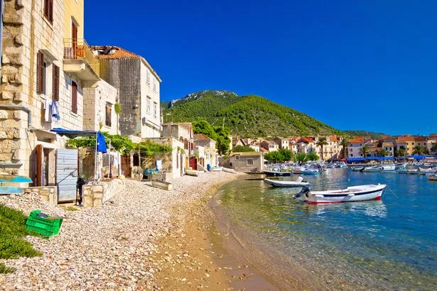 Town of Komiiza beach and old architecture, island of Vis, Croatia. Small boats are visible on the water, with green hills in the distance.