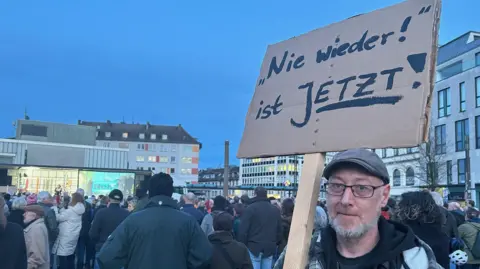 A man with a beard and a flat cap holds up a sign in German that reads 