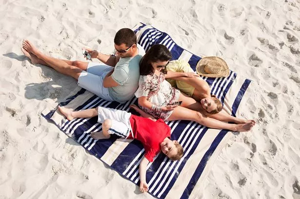 Photo of a young family of four sitting on a large towel at a beach. Husband and his wife sit with their backs against each other while their young son and daughter lie in laughter on either side of them.